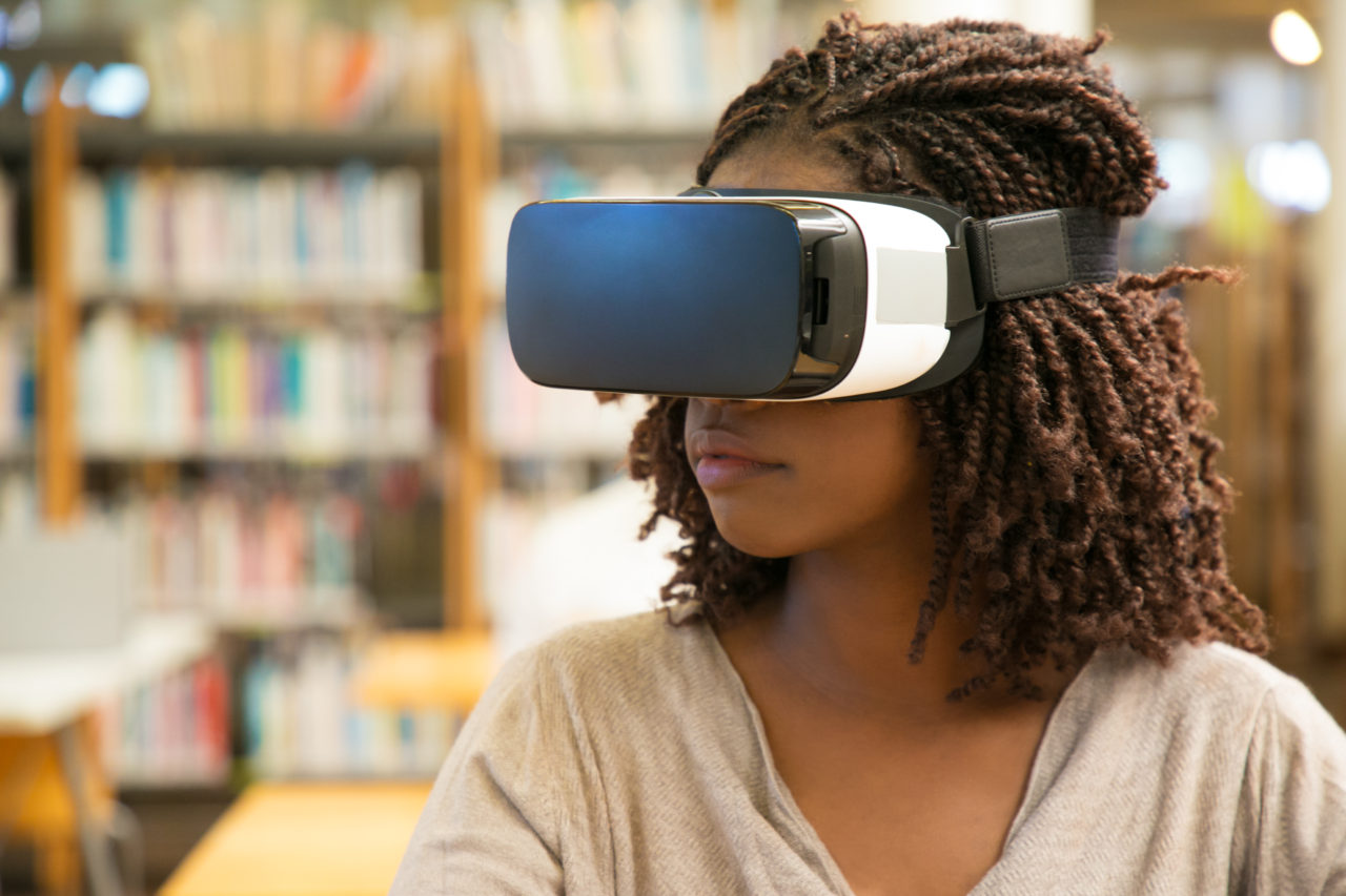 African American student girl using VR glasses for work in library. Young black woman wearing virtual reality headset, sitting at desk. VR technology concept